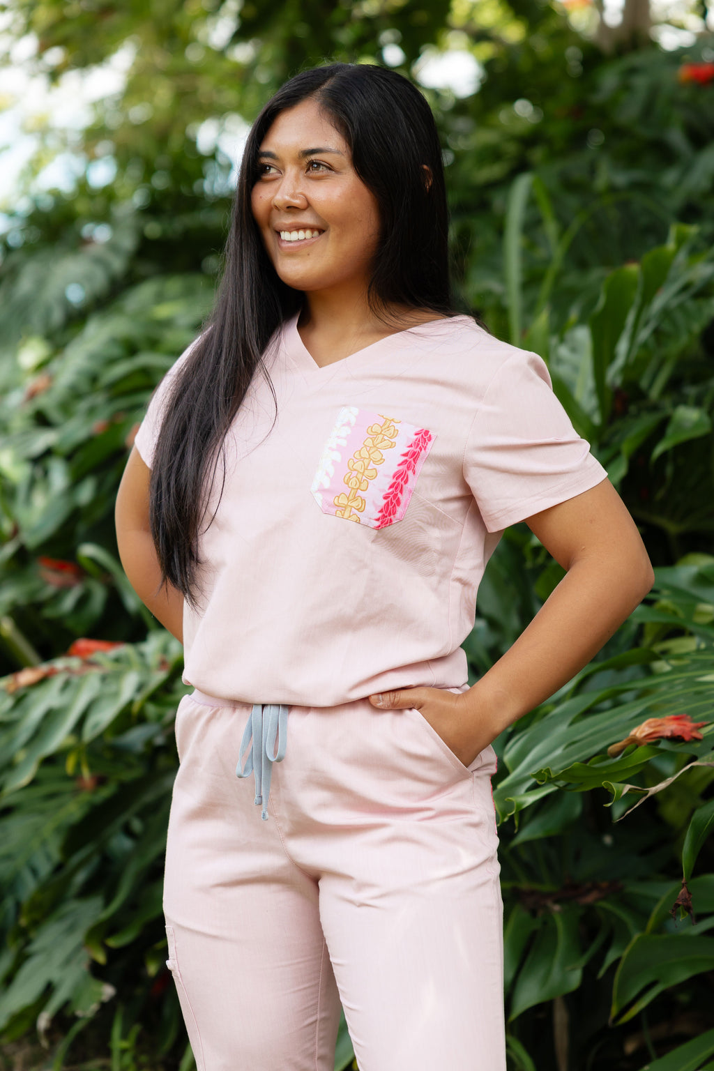 Woman wearing a light pink medical scrub outfit with colorful Hawaii-inspired floral design, standing in front of green foliage.